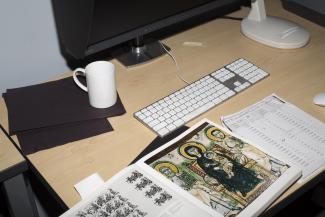 Color photograph of a desk with a computer, keyboard, opened book and coffee mug.